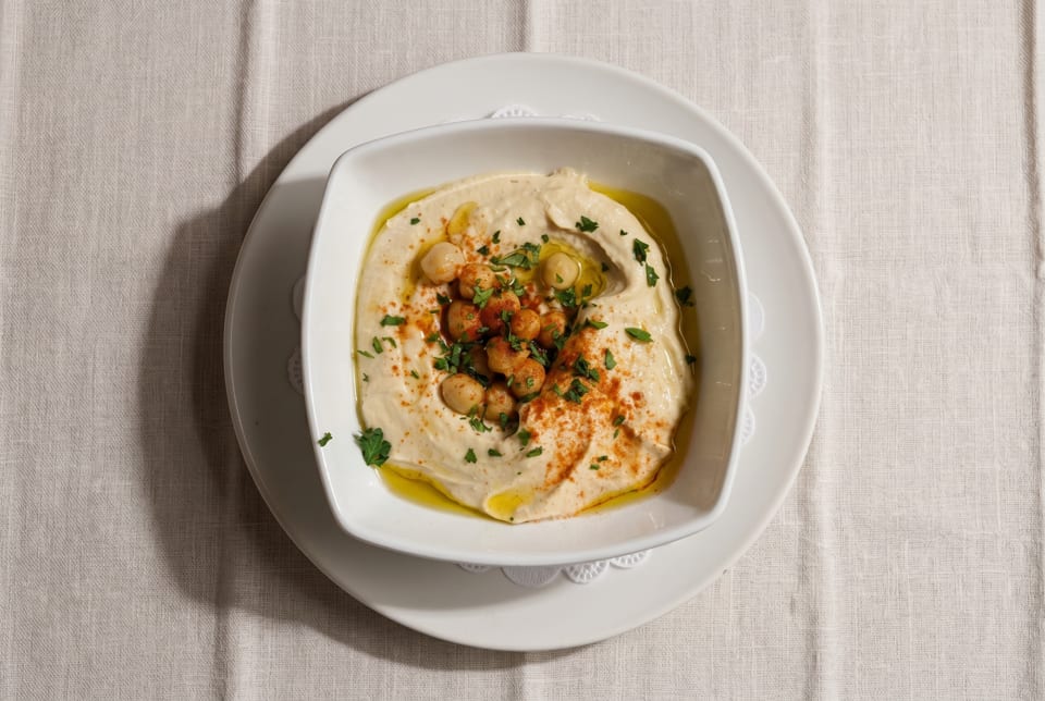 A white bowl of hummus, topped with chickpeas, paprika, and parsley, on a white tablecloth.