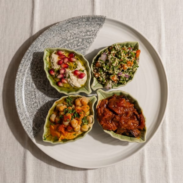 Four leaf-shaped dishes of hummus, tabbouleh, chickpeas, and stewed vegetables on a white plate.