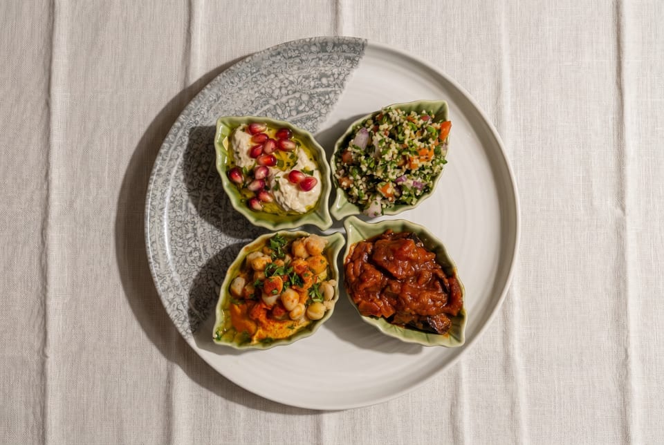 Four leaf-shaped dishes of hummus, tabbouleh, chickpeas, and stewed vegetables on a white plate.